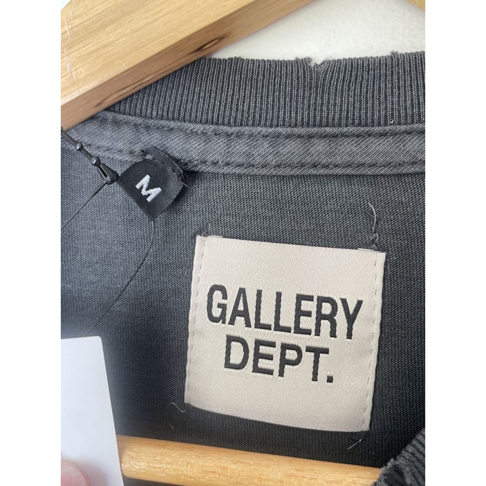 Close-up of a grey Gallery Dept. t-shirt with tags. One tag indicates the size "M". The other tag displays the brand name "GALLERY DEPT." The shirt is hanging on a wooden hanger.