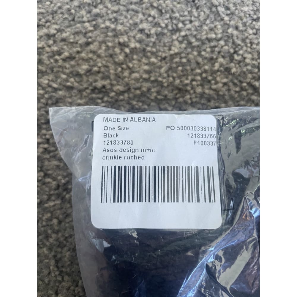 Product tag in plastic wrap. The tag is labeled "MADE IN ALBANIA", "One Size", "Black", "Asos design m+m crinkle ruched", and also includes a barcode and several numbers.