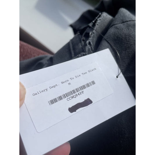 Close-up of a white clothing tag attached to a black garment, with the text "Gallery Dept. Born To Die Tee Black" and the size "M" printed on it. The tag also has a barcode labeled "CCWQ84PF," with a portion of it blacked out.