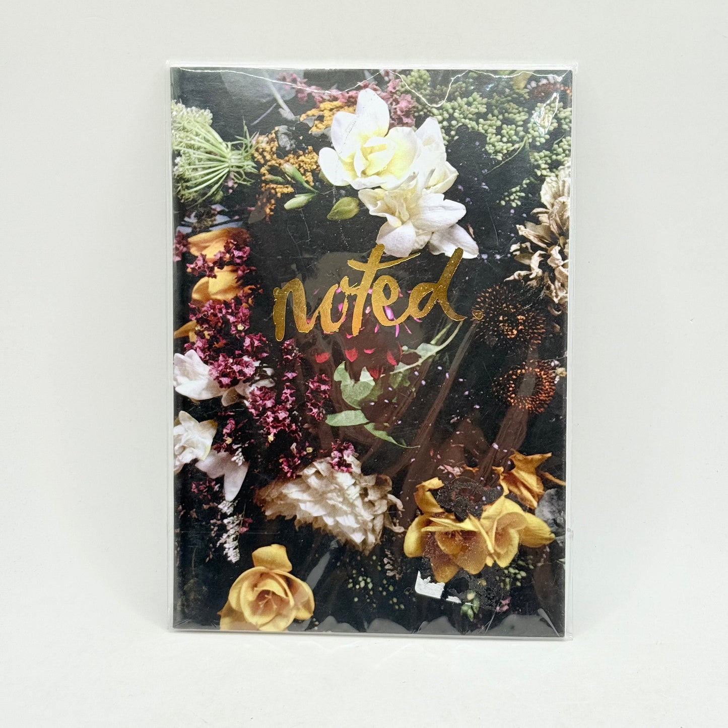 A dark-colored notebook with a floral design and the word ’noted’ in gold script.