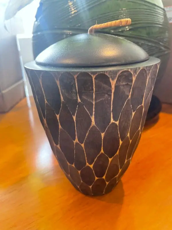 A dark, carved wooden vessel with a domed lid and a curved handle.