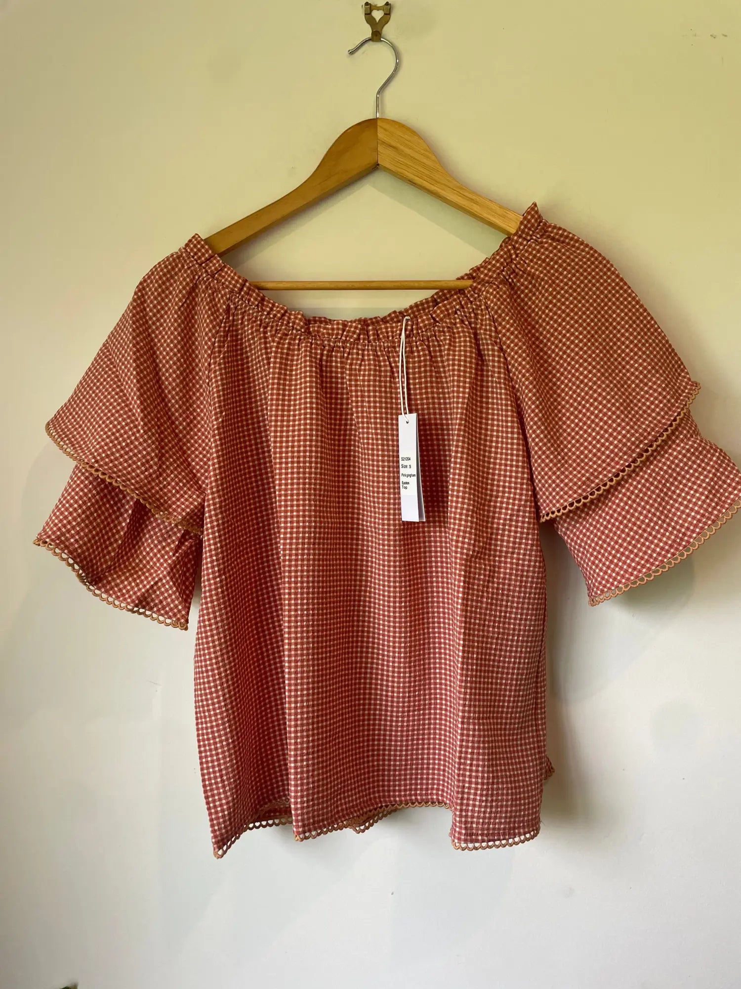 An off-the-shoulder, short-sleeved top with ruffle details on the sleeves and a small gingham pattern in reddish-brown and white.