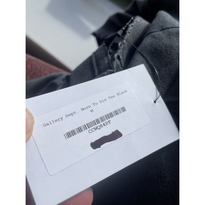 Close-up of a white clothing tag attached to a black garment, with the text "Gallery Dept. Born To Die Tee Black" and the size "M" printed on it. The tag also has a barcode labeled "CCWQ84PF," with a portion of it blacked out.
